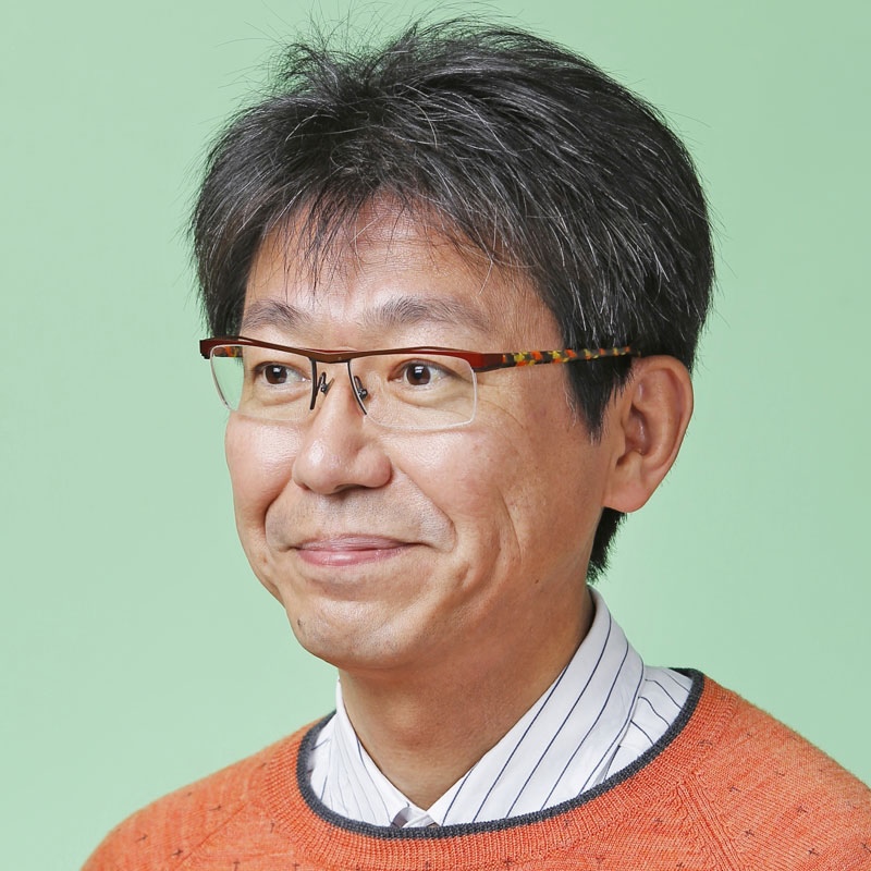 Smiling man with eyeglasses wearing a striped dress shirt under a red sweater.