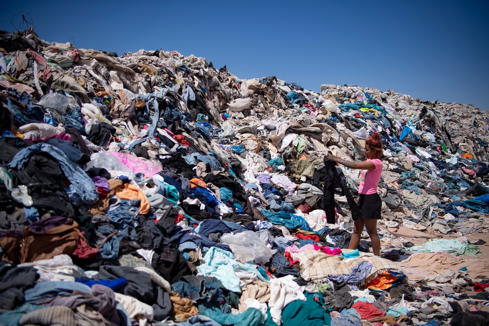 a woman holds up a shirt while standing in a dump with a giant pile of clothes