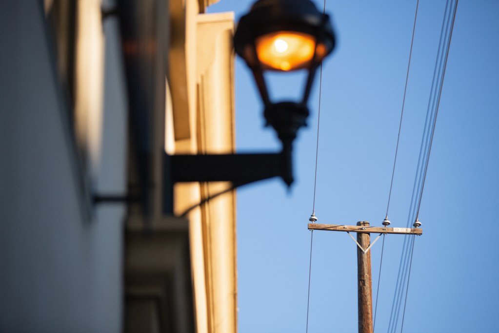 A view of a wooden pole with power lines on a clear blue sky. A lit up lamp outside a home can be seen in the foreground.