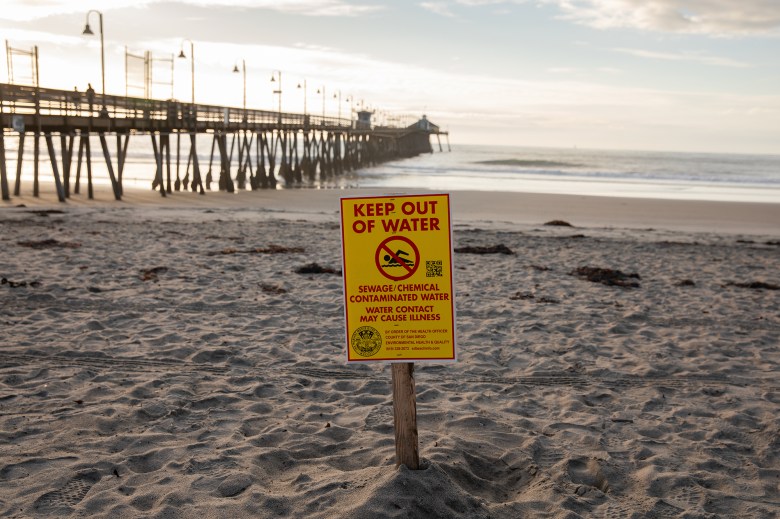 A yellow and red warning sign about sewage and chemical contamination is posted along the shore of a beach. A pier can be seen in the background as the sun begins to set.