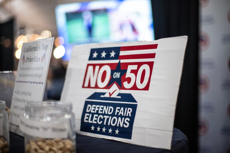 A red, white, and blue sign reading “No on 50: Defend Fair Elections” sits on a table beside jars filled with pistachios and a small display about California’s political priorities. The background shows blurred lights and a screen displaying a broadcast.