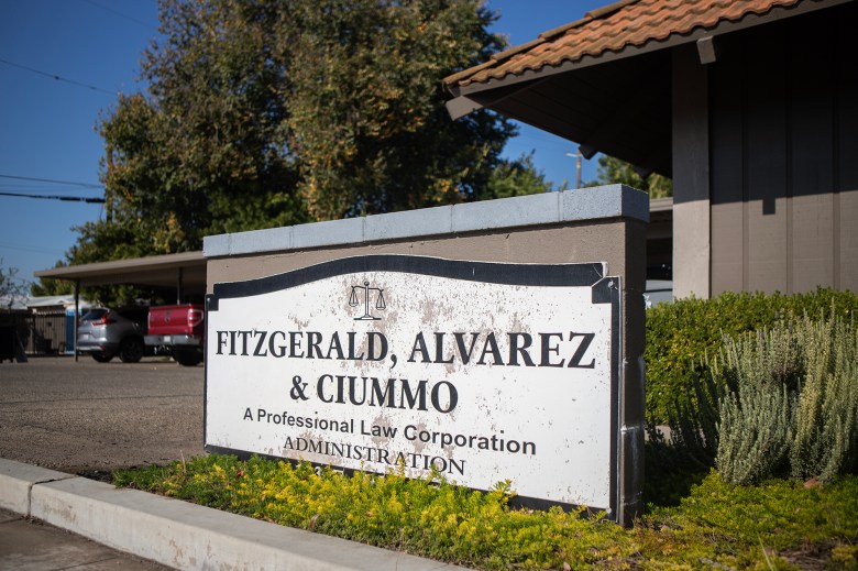 A worn outdoor sign for a law office reads “Fitzgerald, Alvarez & Ciummo – A Professional Law Corporation, Administration,” mounted in front of a low building with bushes and a small parking area in the background.