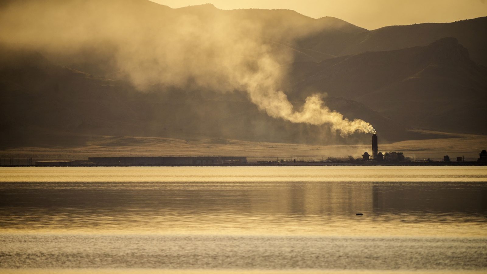 A smokestack billows pollution over a lake with mountains in the background
