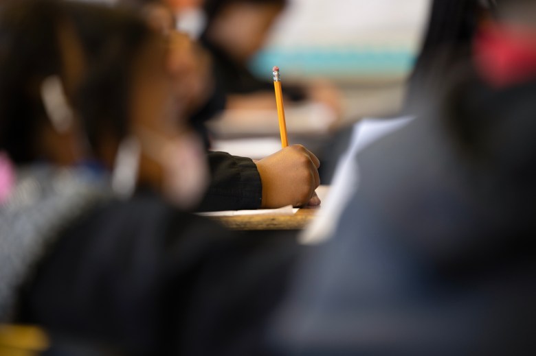 A close-up of a student’s hand holding a pencil above a desk while writing on paper, with other blurred students seated nearby in a classroom.