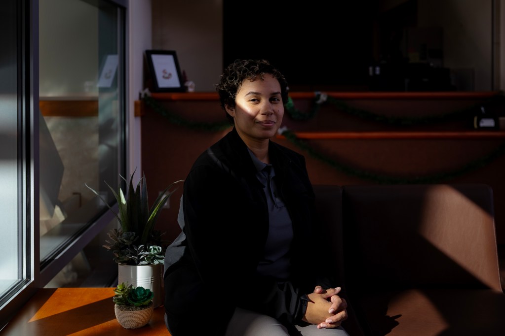 A person with short, curly hair sits indoors near a window, hands clasped in their lap, with a calm expression. Sunlight falls across part of their face and shoulder, while the rest of the room remains in shadow. Small potted plants sit on a ledge beside them, and shelves with framed items and greenery are visible in the softly lit background.