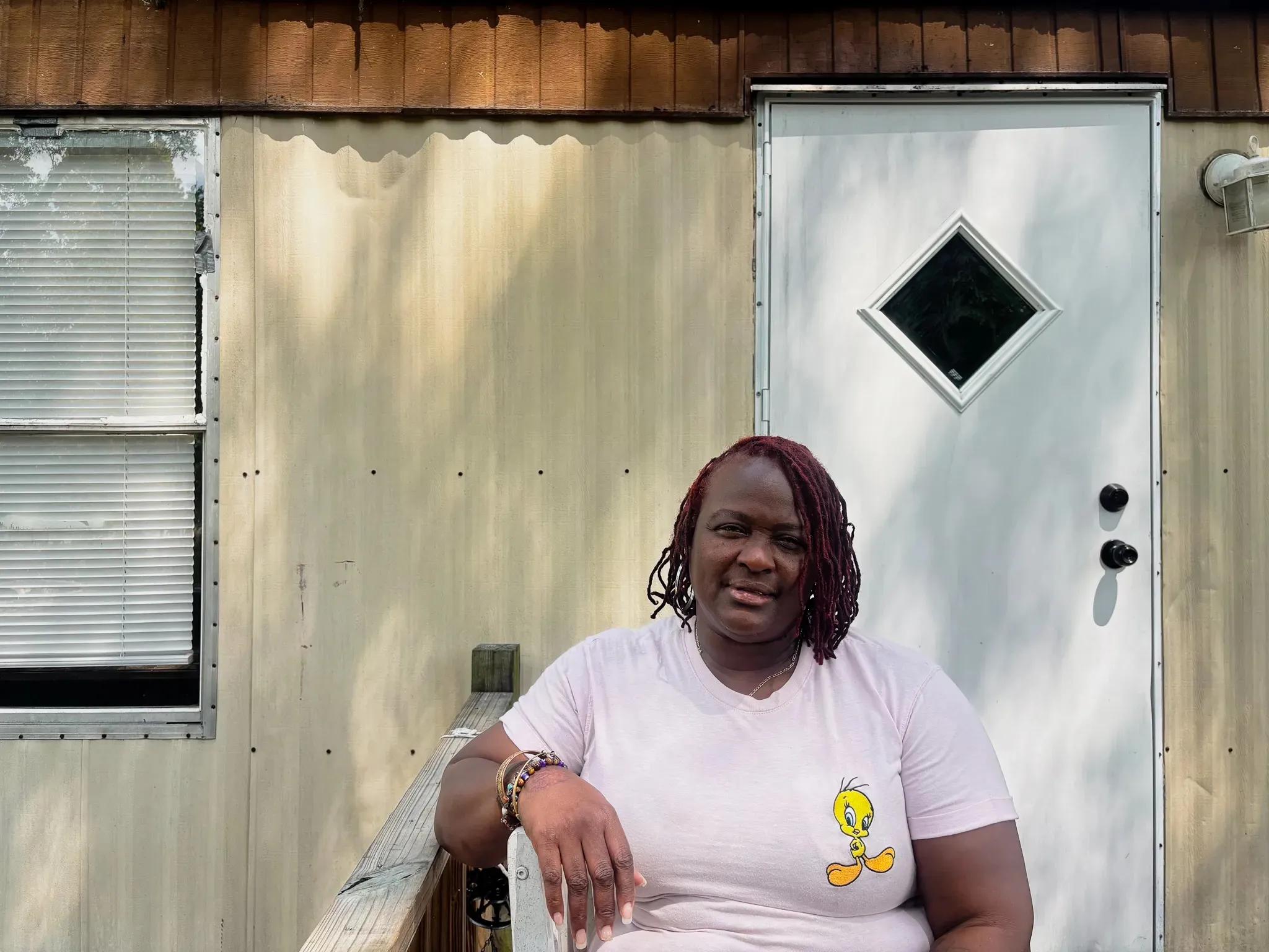 A Black woman in a pink tee shirt sits in a chair outside a house