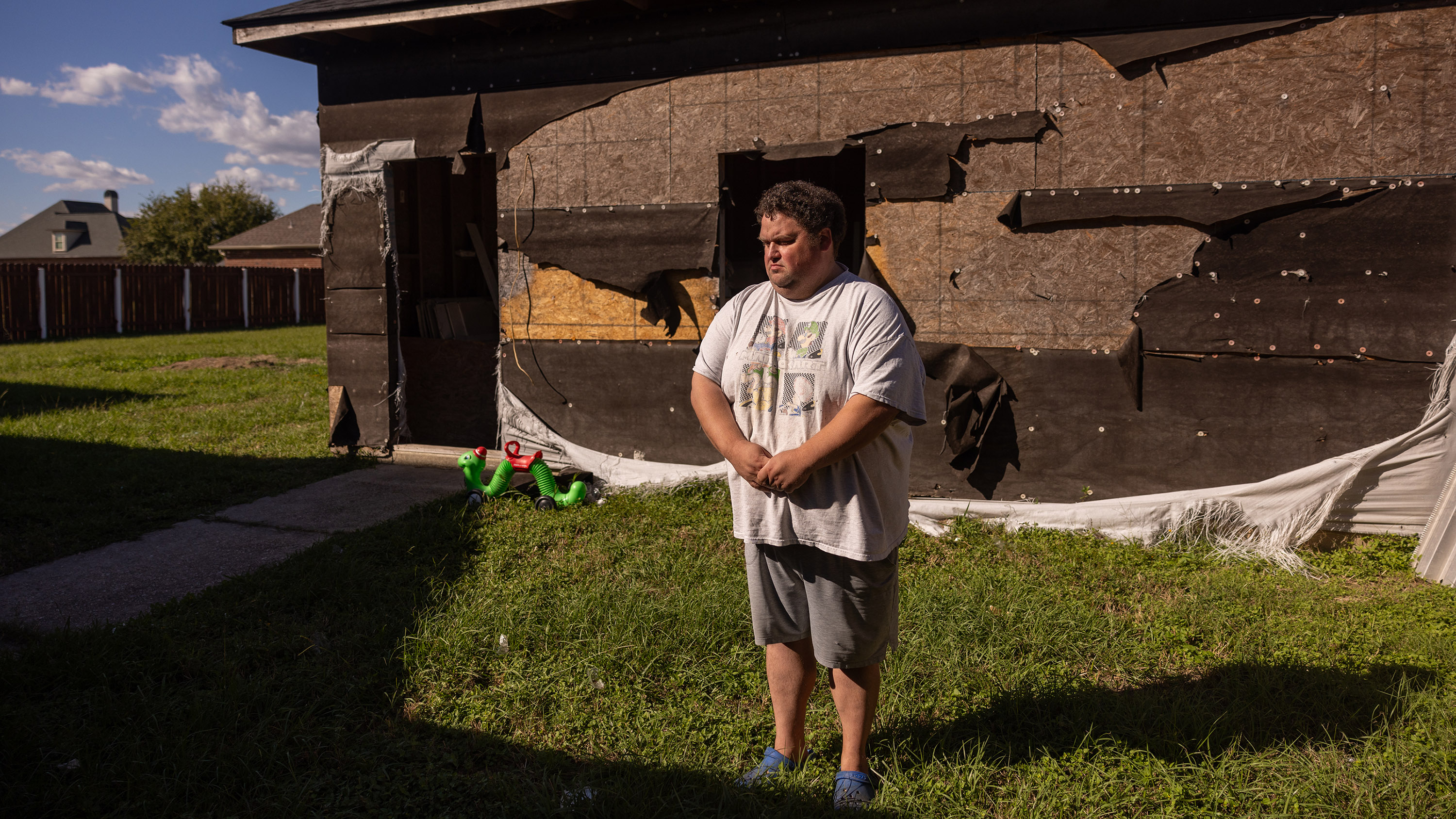 A middle-aged man in a mariokart t-shirt stands in front of a house with a severely damaged facade covered in torn construction tarp and mesh. Near the house, a small green plastic worm toy stands propped up next to a sidewalk
