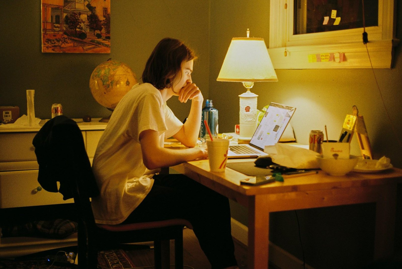 Person sitting at a cluttered desk late at night, staring at a laptop under a lamp.