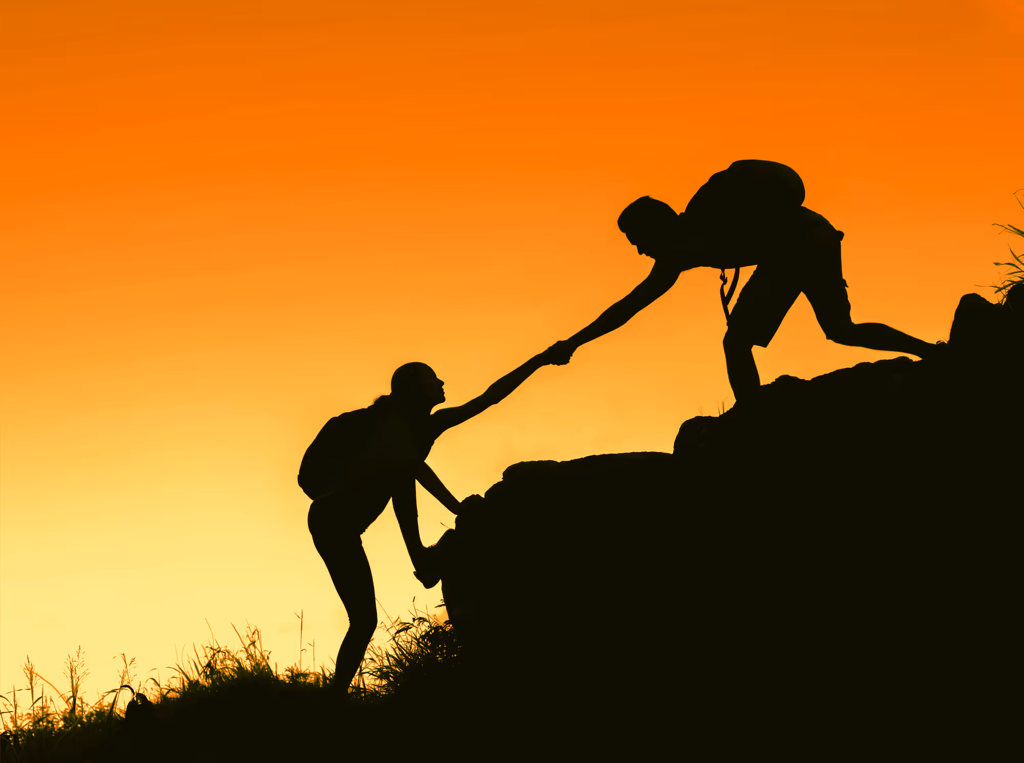 A silhouette of a person helping another climb a hill during a sunset, symbolizing support and teamwork.