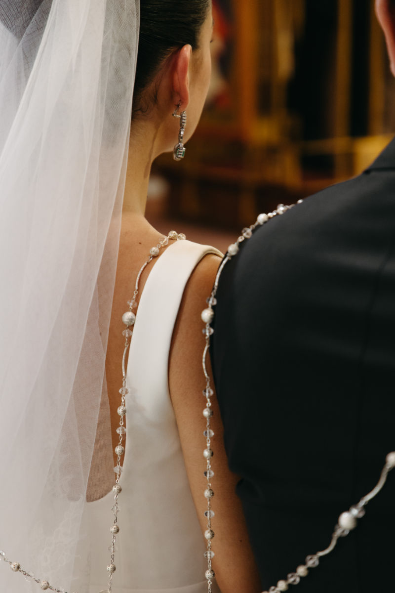 Back view of a bride and groom with decorative pearl and crystal strands draped over their shoulders during a wedding ceremony.