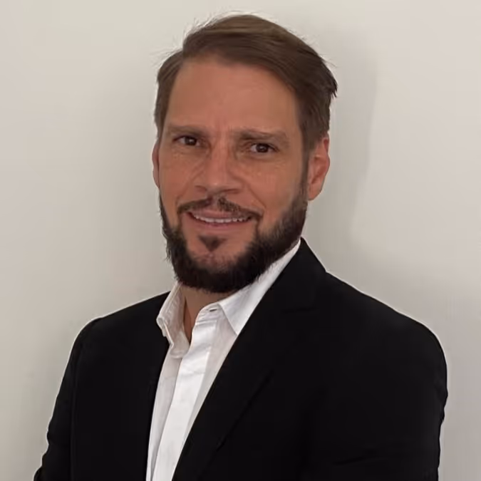 Smiling man with short brown hair and beard wearing a black blazer and white shirt against a plain light background.