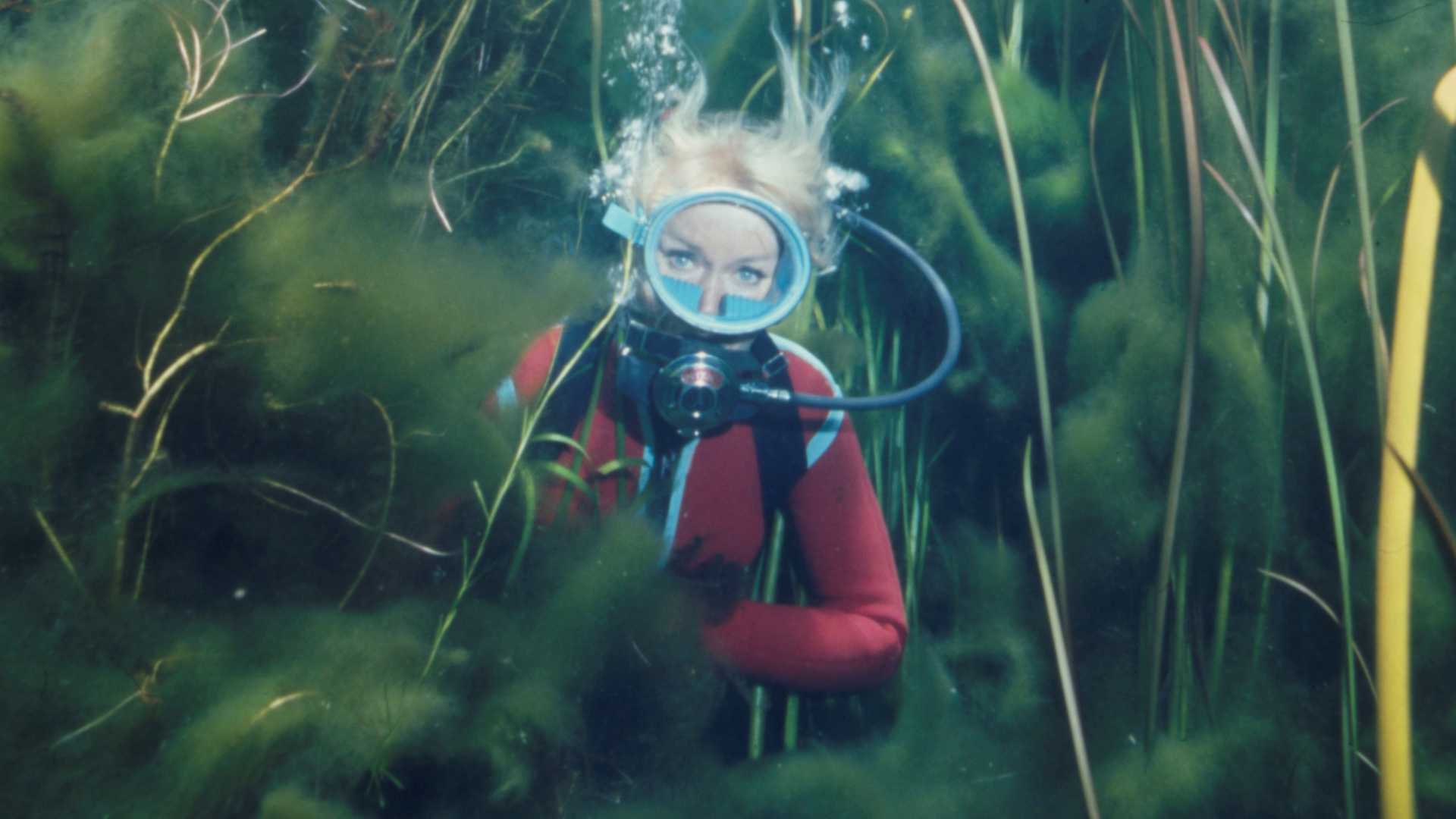 Diver with blonde hair wearing a red wetsuit and blue diving mask underwater among green aquatic plants.
