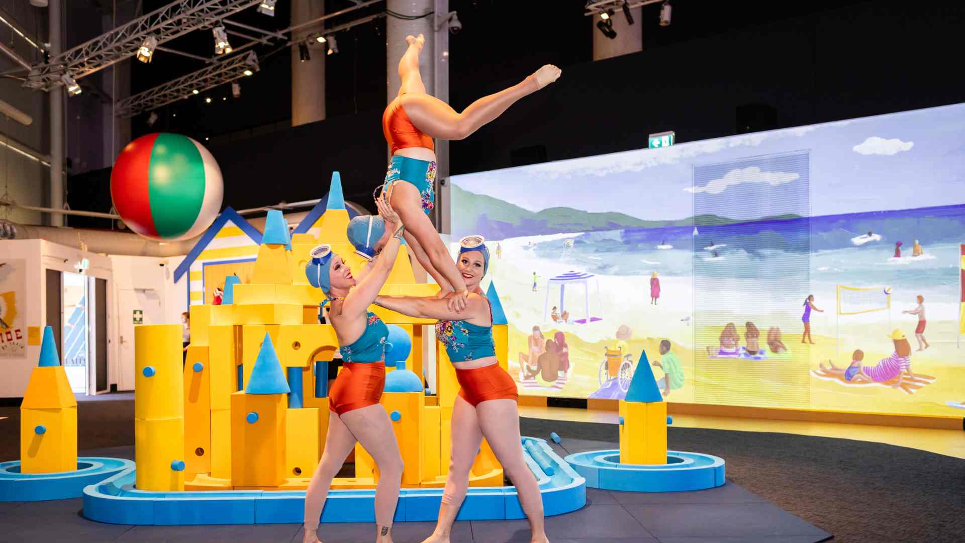 Three women in vintage swimwear performing an acrobatic balancing act in front of a yellow and blue sandcastle structure with a beach mural backdrop.