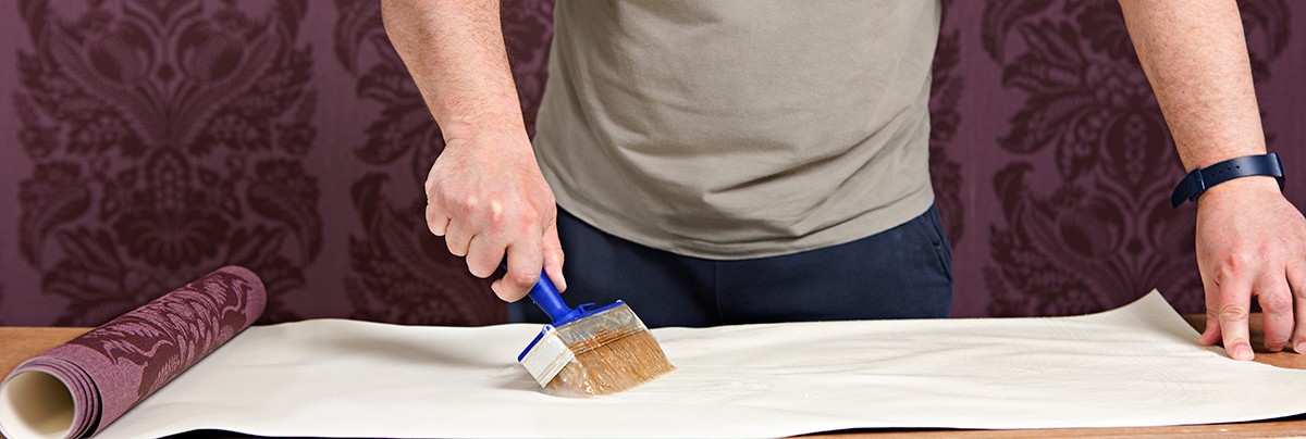 Man placing glue onto the back of purple coloured, flower embossed wallpaper.