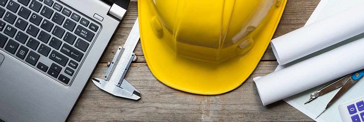 Wooden table with a yellow construction helmet, laptop and plans