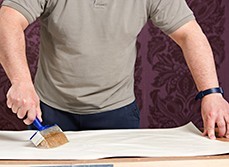 Man placing glue onto the back of purple coloured, flower embossed wallpaper.