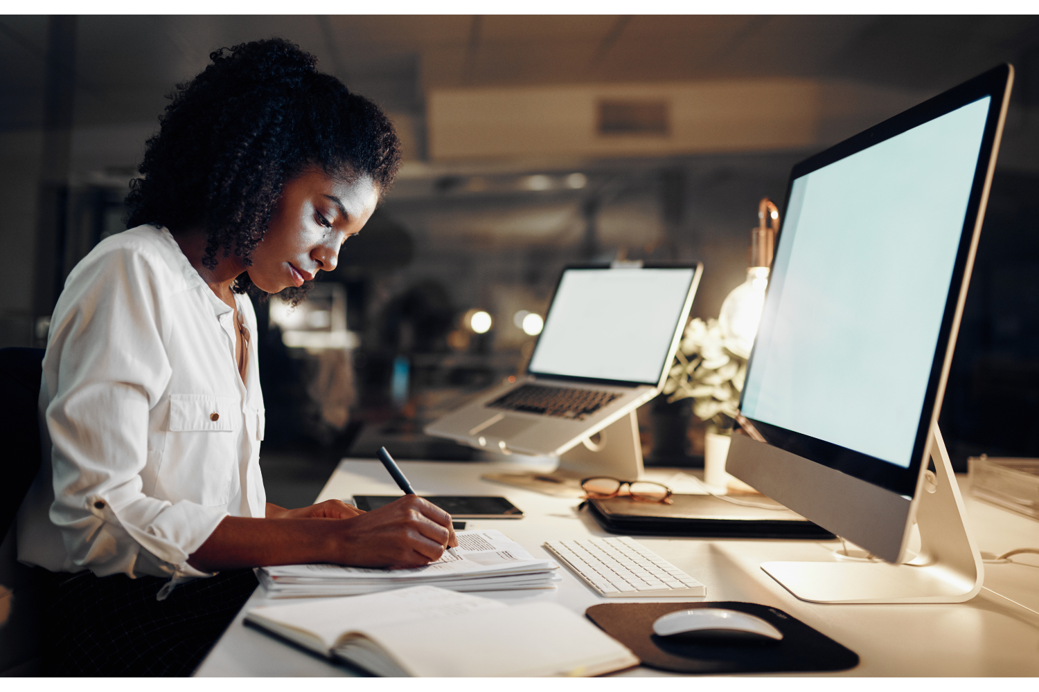 A theatre administrator works at a desk with a laptop and desktop computer, taking notes in a paper document.