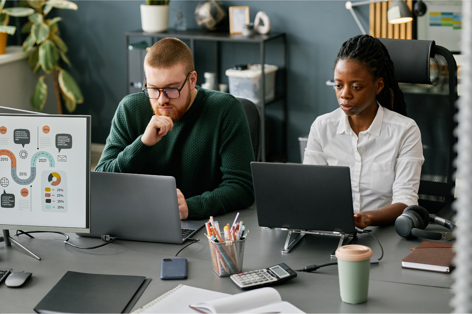 Two theatre administrative staff members work at laptops in an office, reviewing materials and concentrating on multiple tasks at a shared desk.
