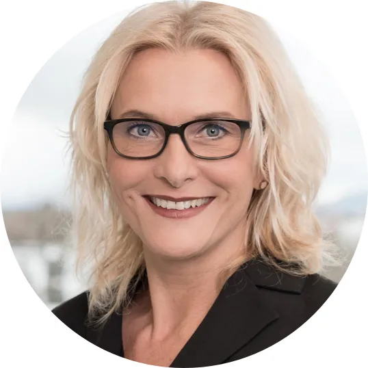 Nahaufnahme einer lächelnden Frau mit blonden Haaren, schwarzer Brille und schwarzem Blazer vor unscharfem Hintergrund.