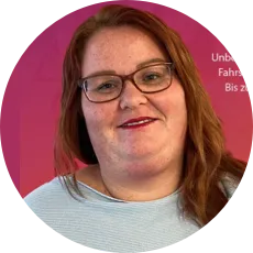 Porträt einer lächelnden Frau mit roten Haaren, Brille und hellblauem Pullover vor einem rosa Hintergrund.
