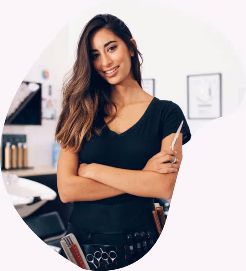 Smiling female hairstylist with arms crossed holding scissors in a salon with tools belt.