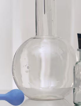 Clear round-bottom laboratory flask beside a small glass bottle and blue rubber dropper on a white surface.