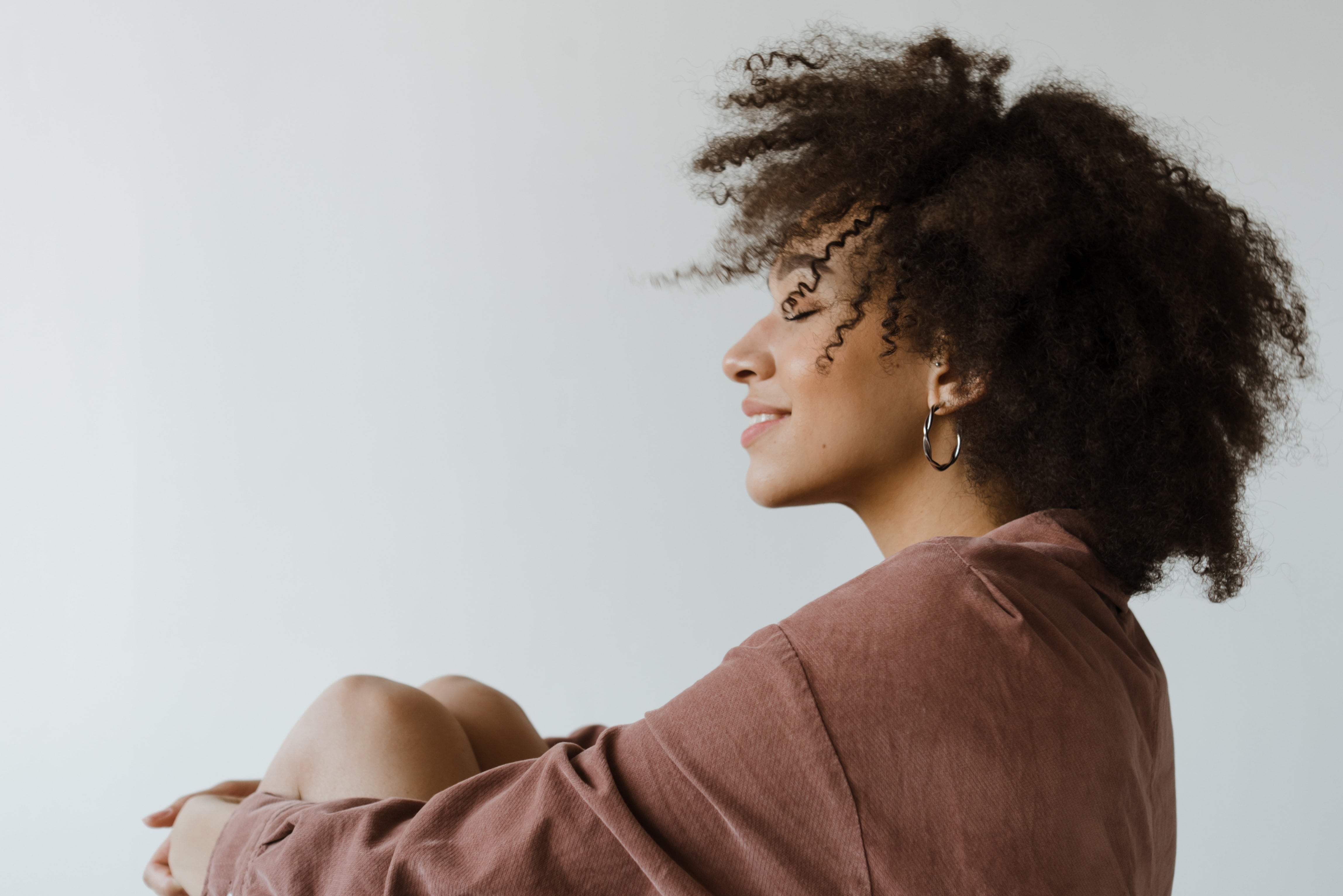 Side profile of a smiling woman with curly hair wearing hoop earrings and a brown shirt against a plain white background.