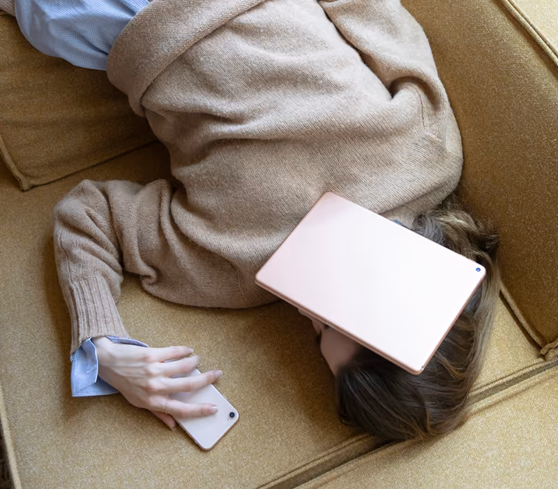 Person lying curled on a mustard couch holding a smartphone with a tablet resting on their face.