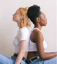 Two women sitting back to back with eyes closed, wearing white tops and jeans against a plain background.