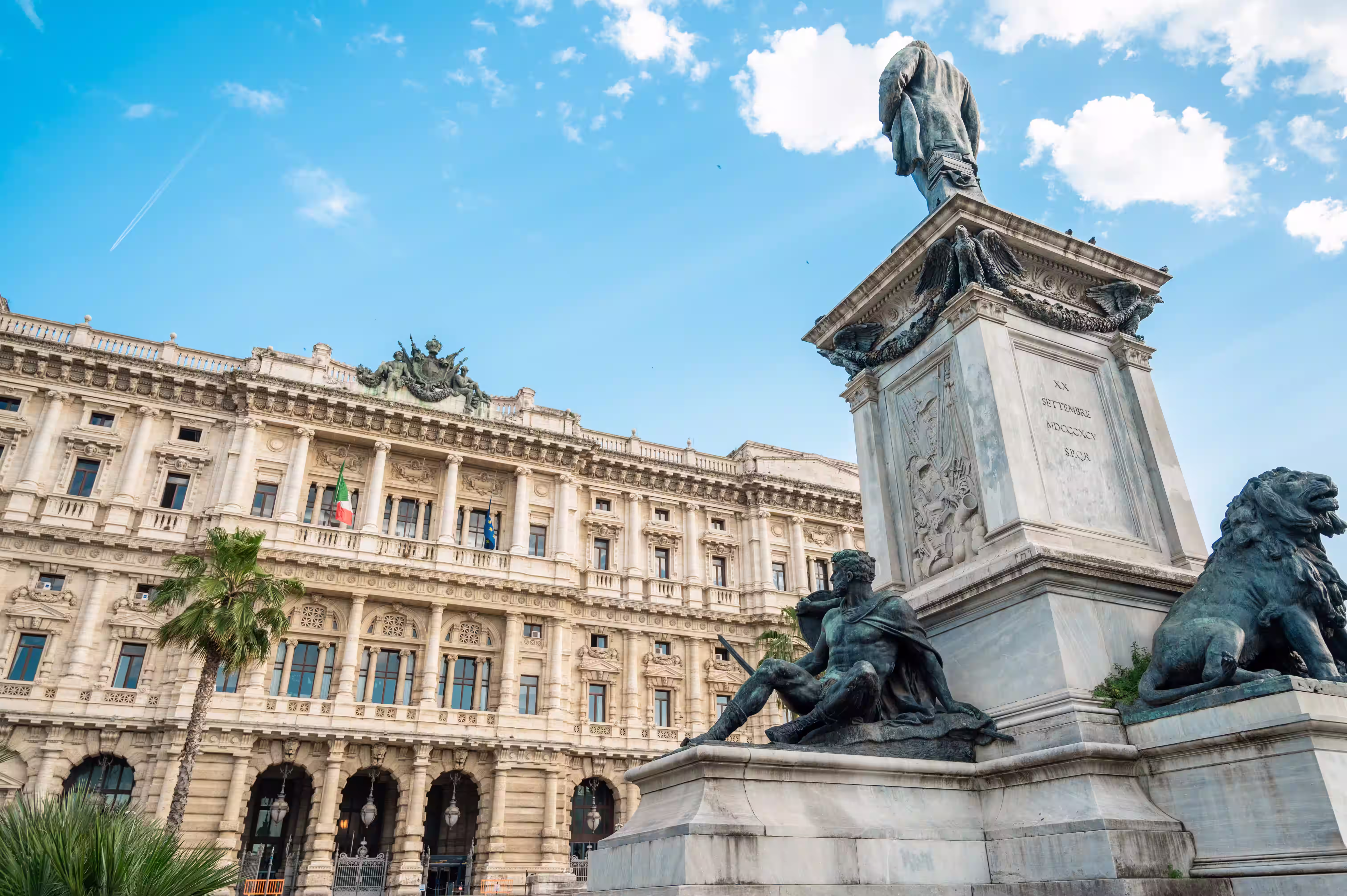 Monument with statues of a seated man and a lion in front of a historic ornate building under a blue sky with light clouds.