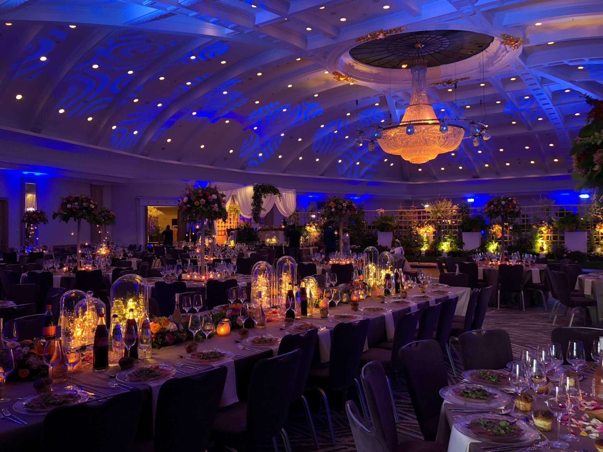 Bijzonder overzicht van een prachtig aangeklede trouwlocatie met grote luster, blauwe plafondverlichting en warme lichtaccenten aan tafel.