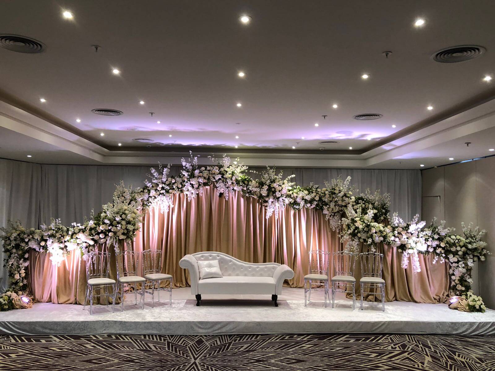 Centraal een luxueuze witte zetel met een achtergrondwand met roze draperieën en bovenaan afgewerkt met bloemen.