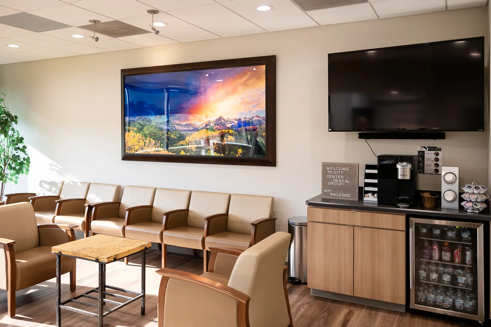 Waiting area with beige armchairs and a small wooden table in front, a large wall-mounted TV, a coffee station with a Keurig machine, and a framed picture of a mountain landscape on the wall.