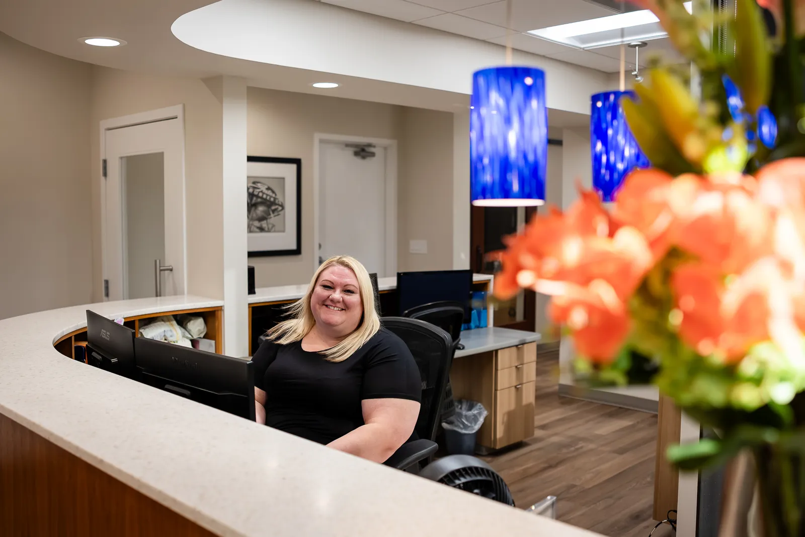 Smiling woman with blonde hair sitting behind a curved reception desk with dual monitors in a modern office interior featuring blue pendant lights and wooden flooring.