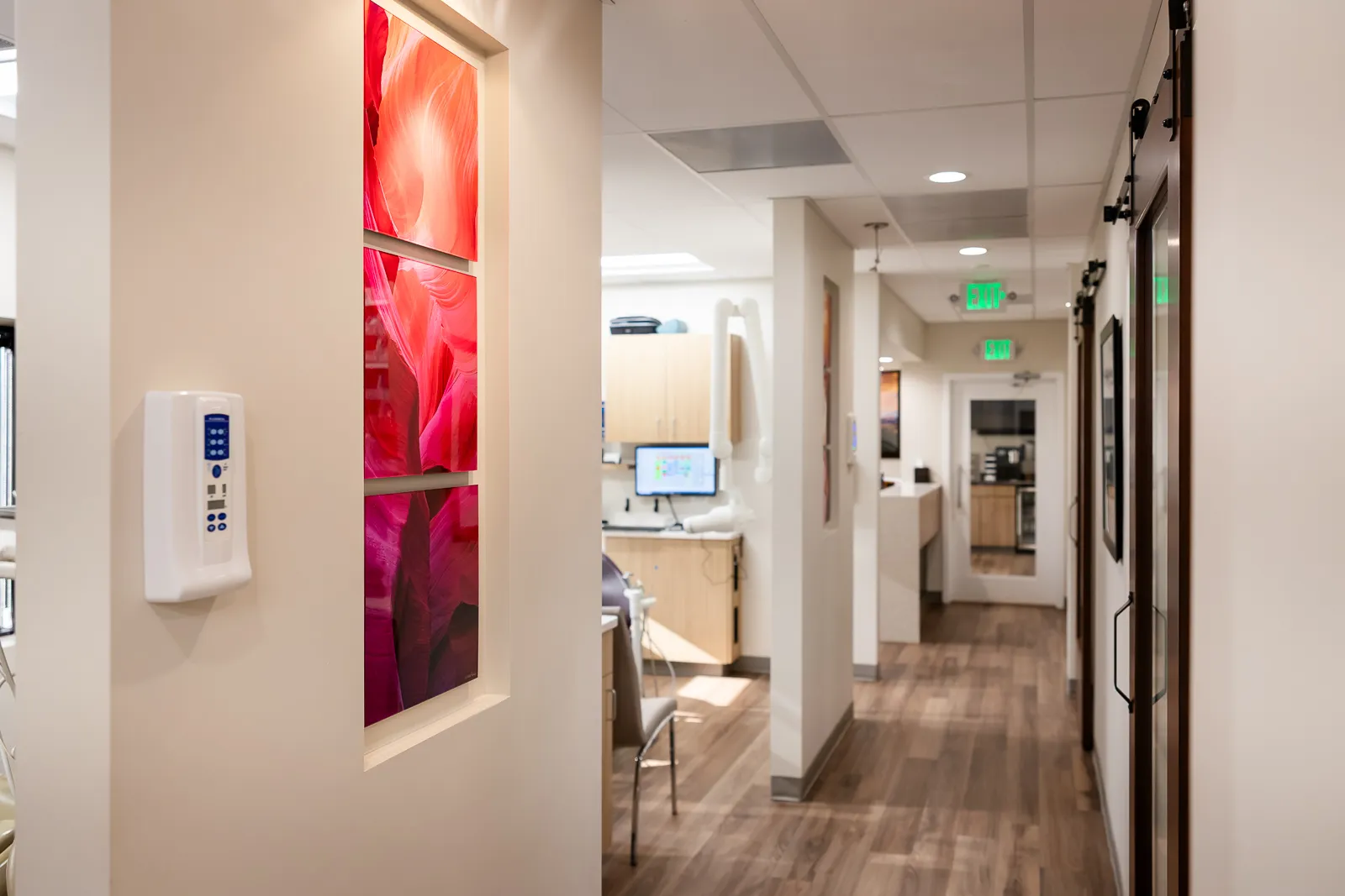 Interior hallway of a modern dental office with wood-look flooring, beige walls, and ceiling with recessed lighting. There is a mounted white medical device on the left wall next to a decorative inset with three red and pink abstract art panels. A dental chair and workstation with computer monitor are visible in a side room. The hallway leads to a white door with glass panel and exit signs above.
