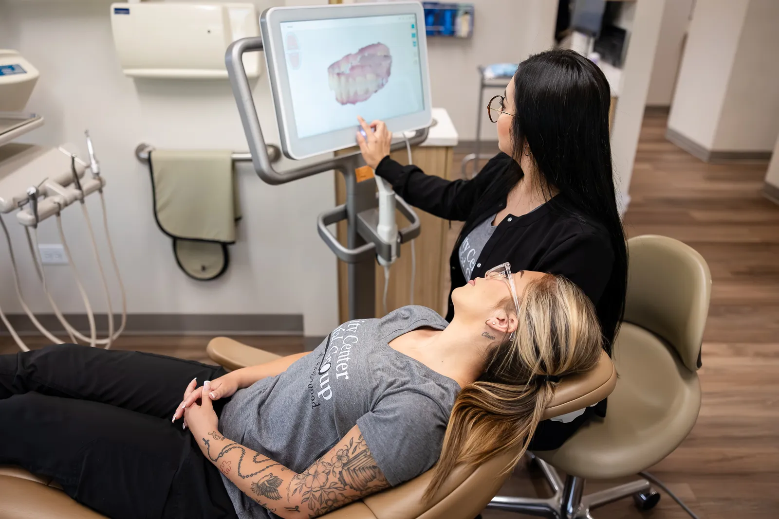 A dental professional wearing glasses and black clothing shows a digital 3D image of a set of teeth on a screen to a patient reclining in a dental chair with tattoos on her arm and wearing a gray t-shirt.