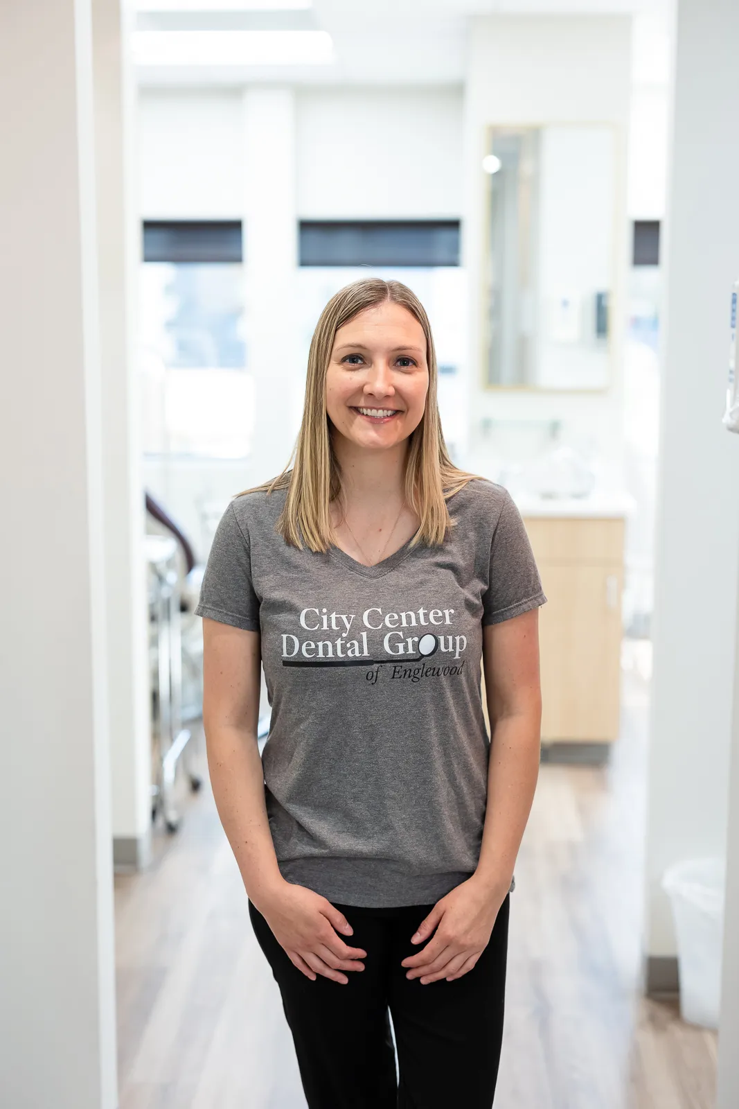 Smiling woman with straight blonde hair wearing a gray City Center Dental Group of Englewood t-shirt standing in a bright dental office hallway.