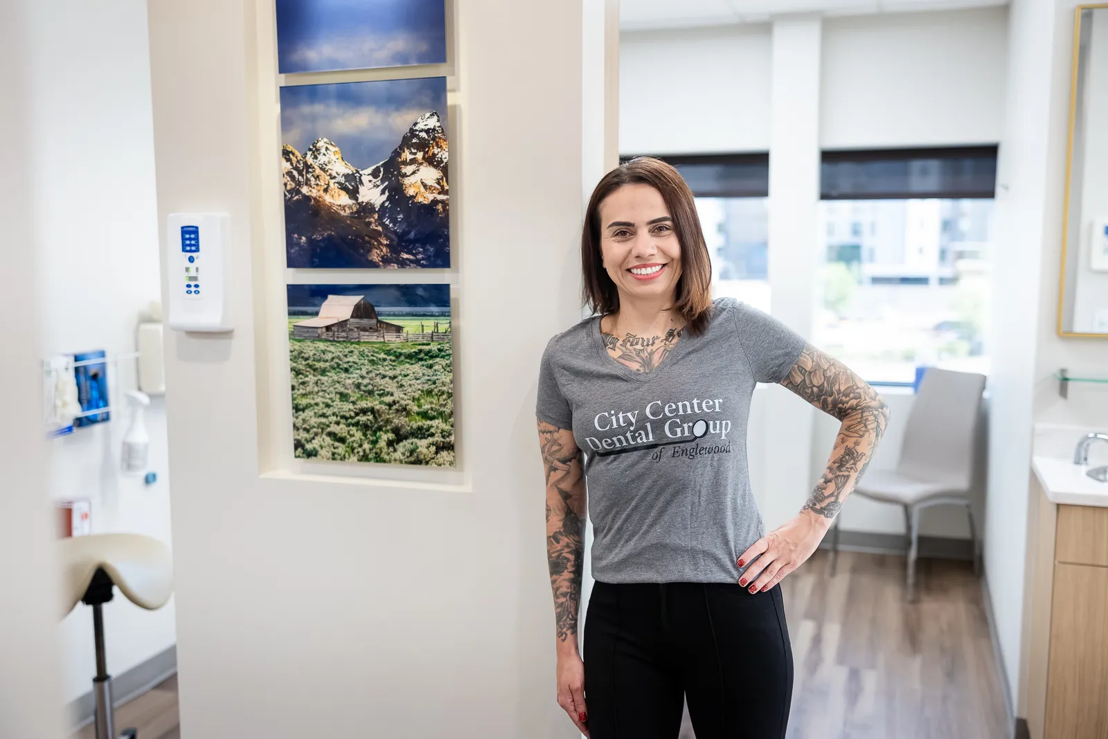 Smiling woman with tattoos on her arms and chest wearing a gray t-shirt that reads 'City Center Dental Group of Englewood' standing in a dental office hallway with framed photos of mountains and a barn on the wall behind her.