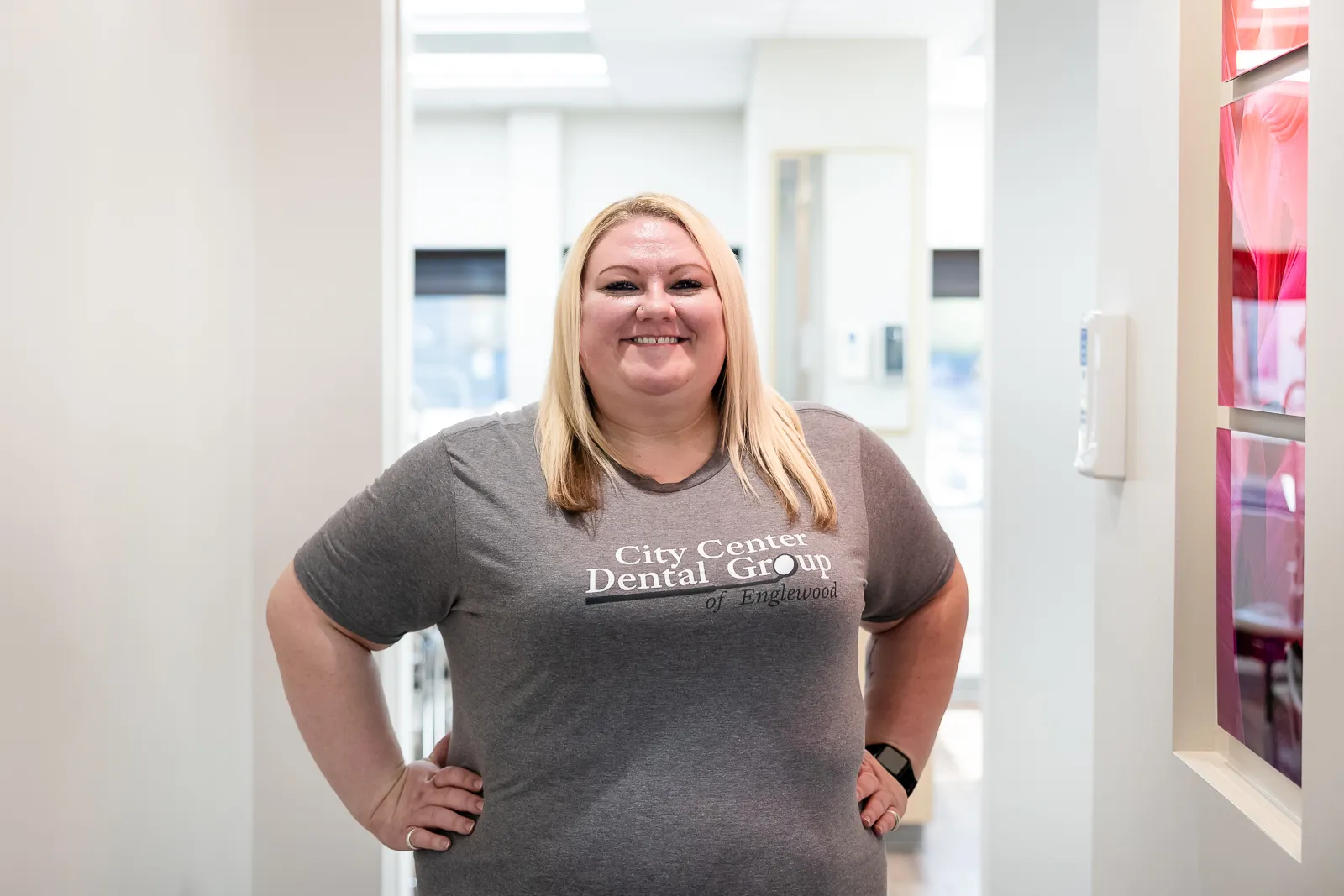 Smiling woman with blonde hair wearing a gray City Center Dental Group of Englewood t-shirt standing in a bright hallway with her hands on her hips.