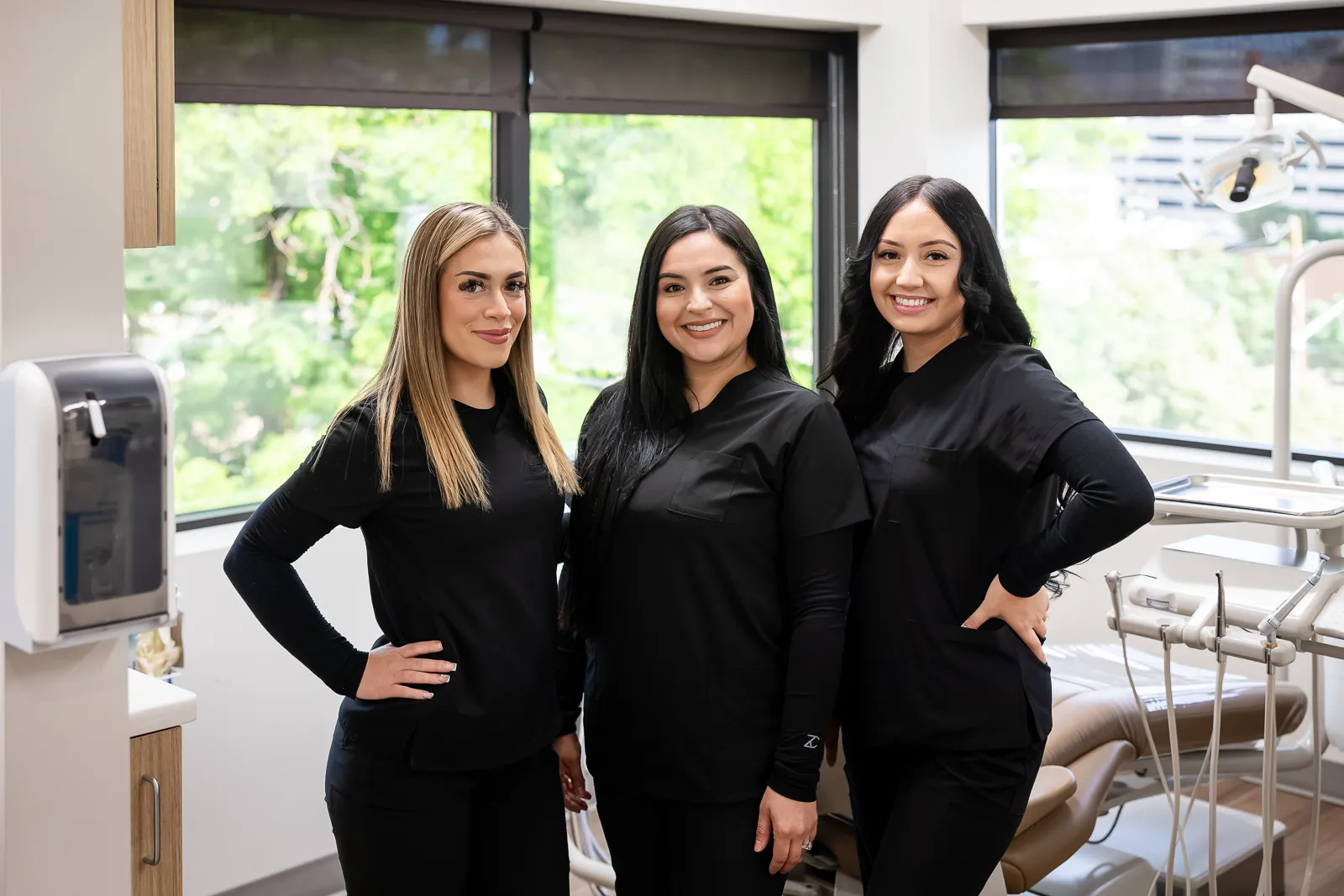 Three dental professionals wearing black scrubs standing side by side with hands on hips, smiling in a bright dental office with large windows and dental chairs visible in the background.