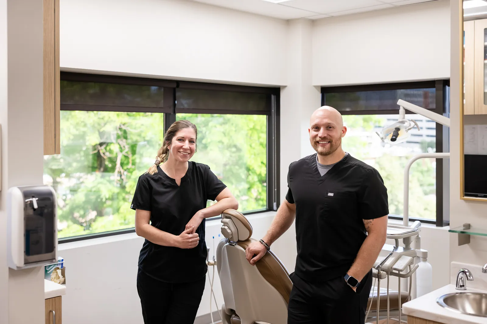 A smiling female and male dentist stand beside a dental chair in a bright dental office with large windows and green trees visible outside.