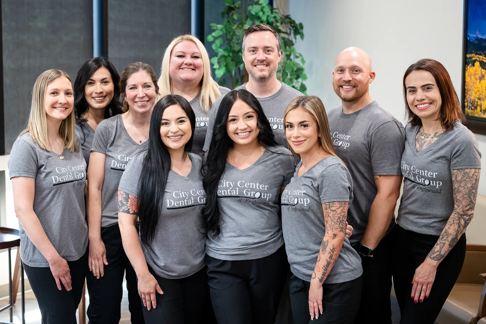 Group of ten smiling dental office staff wearing matching grey City Center Dental Group t-shirts, standing closely together indoors.