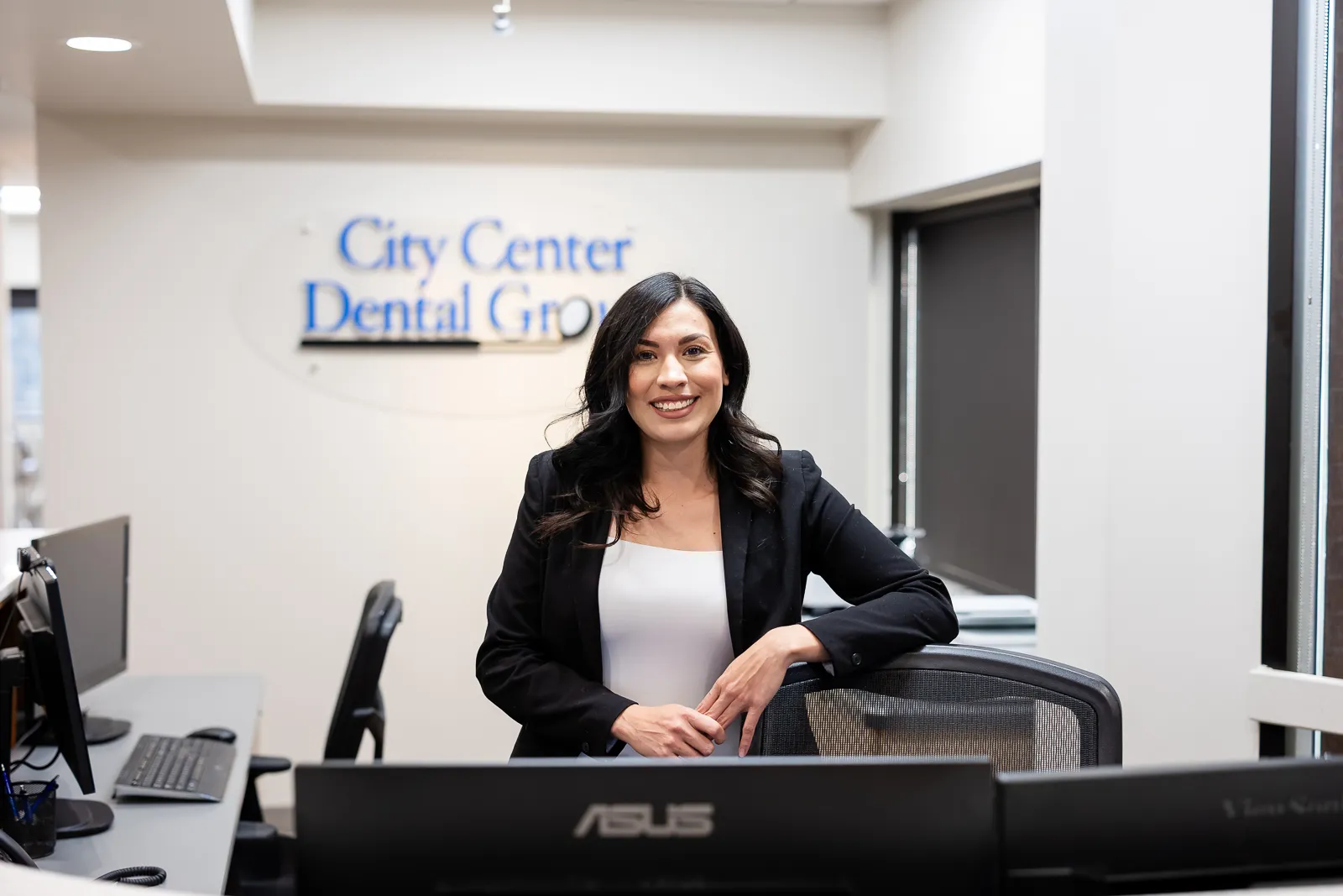 A woman with dark hair wearing a black blazer and white shirt stands smiling behind a desk in an office with computer monitors, with a City Center Dental Group sign blurred in the background.