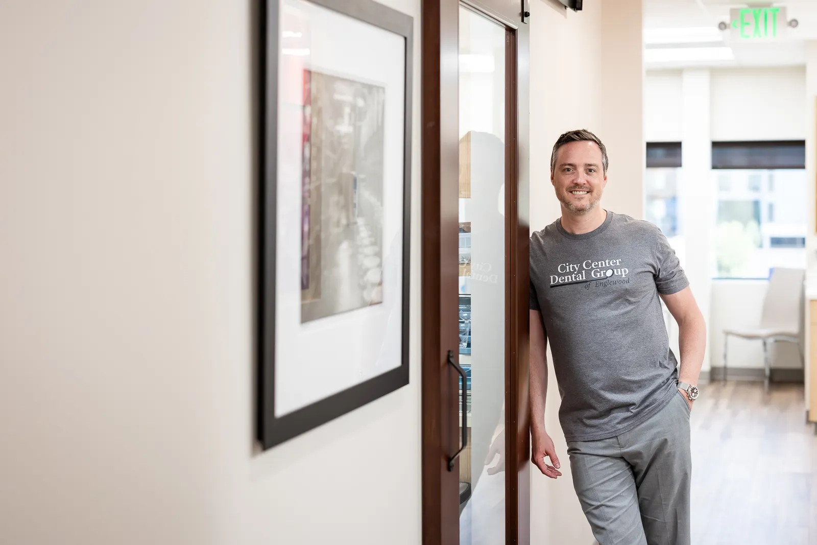 A man wearing a gray City Center Dental Group of Englewood t-shirt stands smiling and leaning against a glass door in a bright, modern office hallway with light walls, wood flooring, and framed wall art nearby.