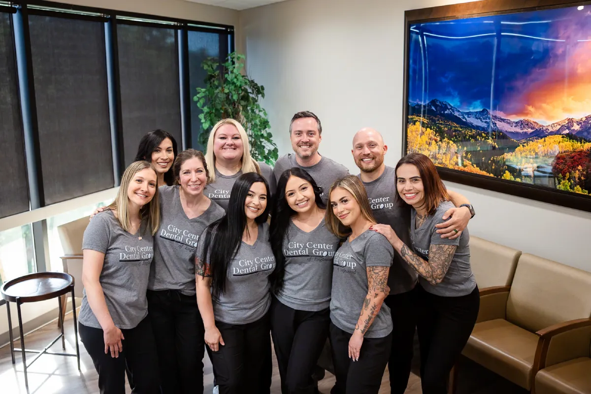 Group of nine smiling dental professionals wearing matching grey City Center Dental Group t-shirts posing indoors in a brightly lit room with windows, plants, and a colorful landscape painting on the wall.