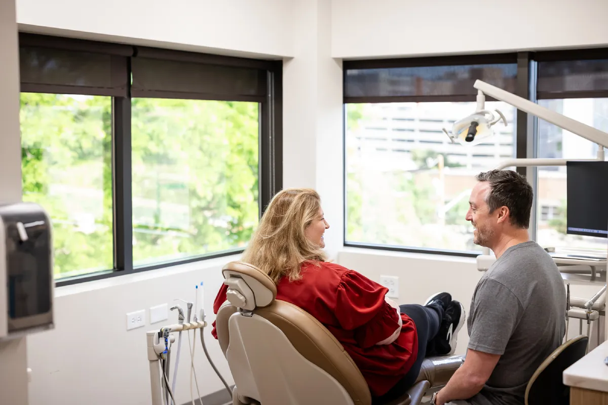 A woman in a red blouse sits relaxed in a dental chair facing a man in a gray t-shirt who smiles at her in a bright dental office with large windows showing green trees outside.