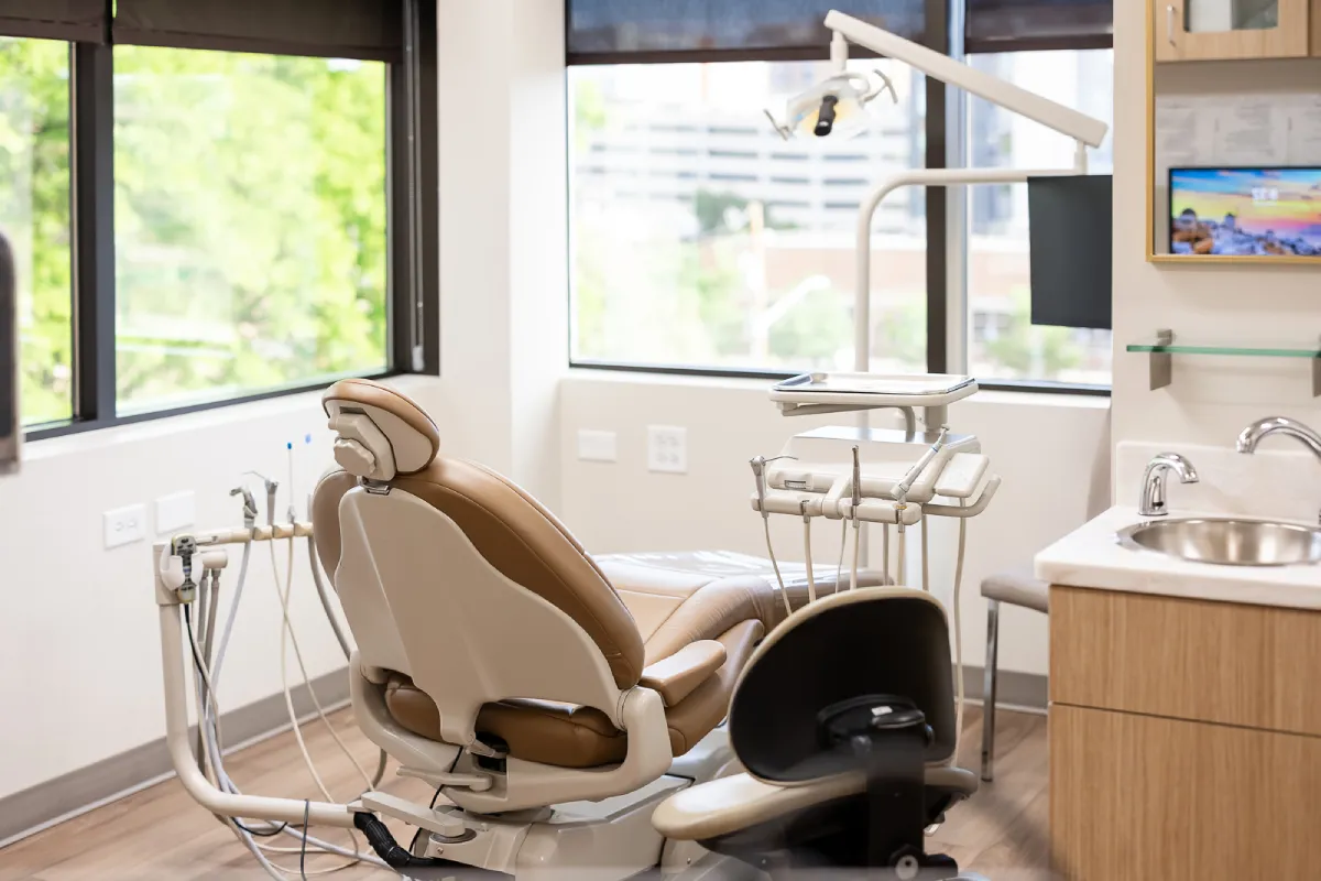 Modern dental treatment room with a brown and beige dental chair, dental tools, overhead light, large windows with green outdoor view, wood floor, sink, and mounted TV screen.