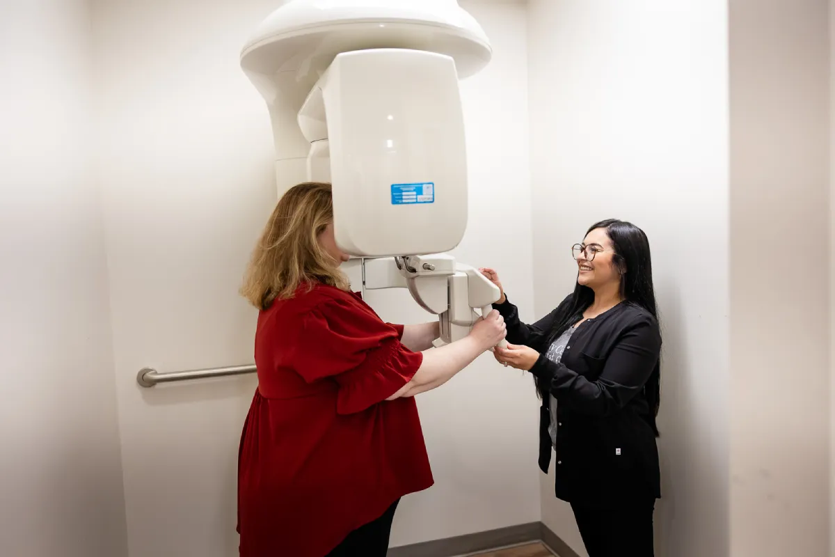 A dental professional assists a patient standing at a panoramic dental X-ray machine in a small room with white walls and a metal grab bar on the wall.