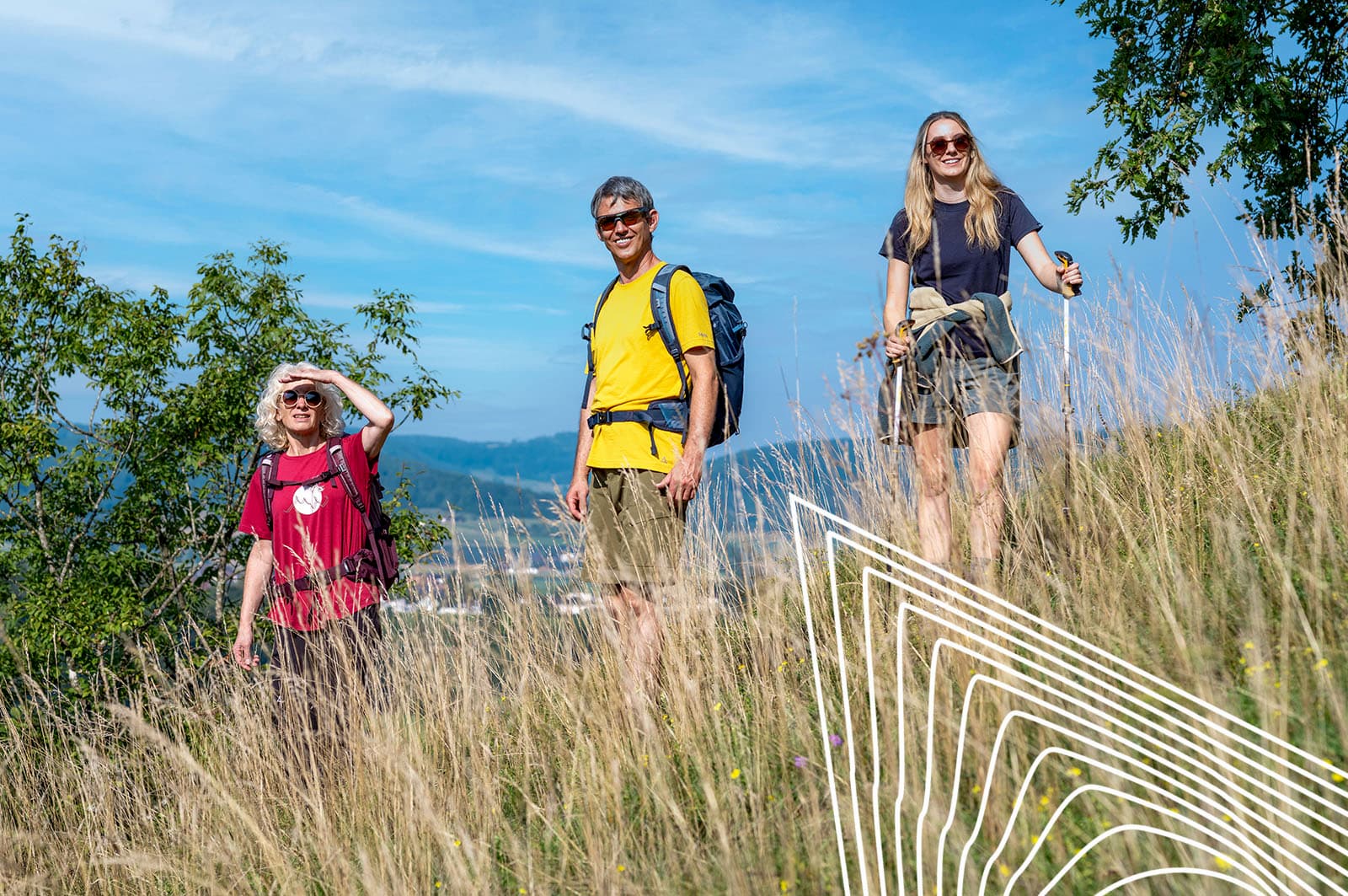 Gruppe von drei Wandernden in Sommerkleidung steht in hohem Gras auf einem Hügel. Alle tragen Sonnenbrillen und Rucksäcke, eine Person hält Wanderstöcke. Im Hintergrund sind Bäume und ein weiter Blick ins Tal unter blauem Himmel zu sehen.