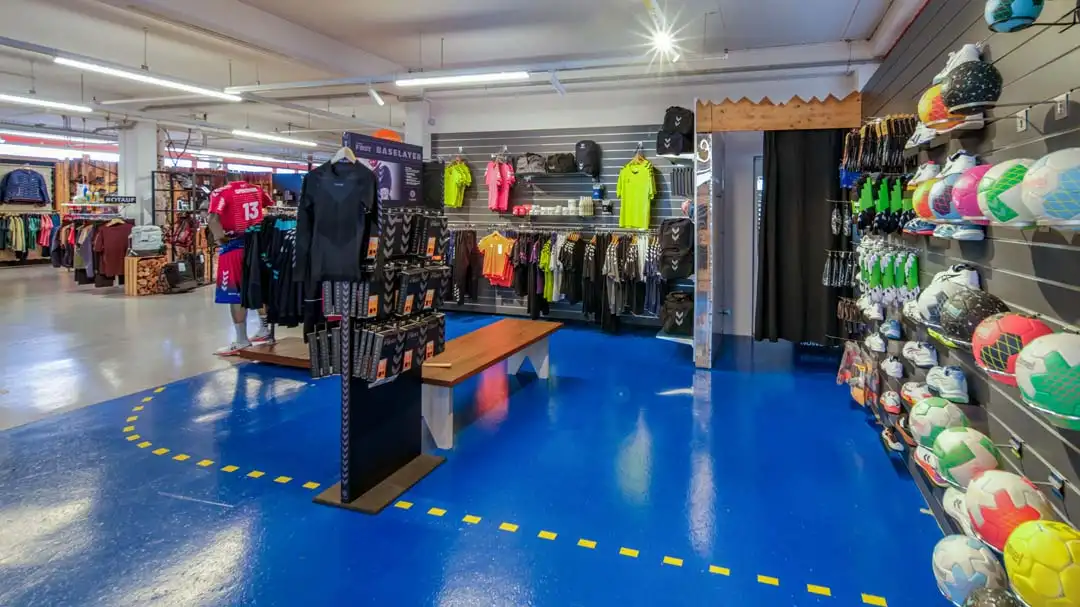 Sportgeschäft mit blauem Boden, Regalen voller Sportbälle, Trikots und Sportbekleidung.